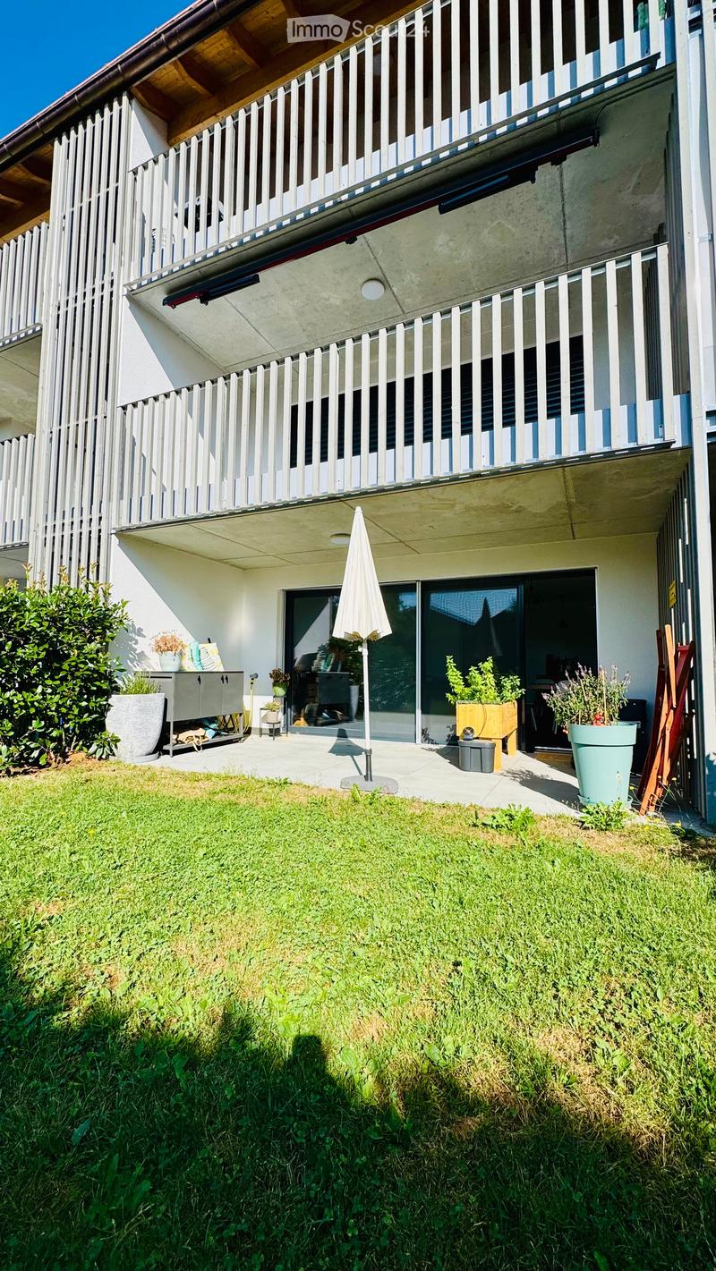 Garden area with a grassy lawn, a small patio with potted plants, an umbrella, and a modern building with balconies and railings in the background.