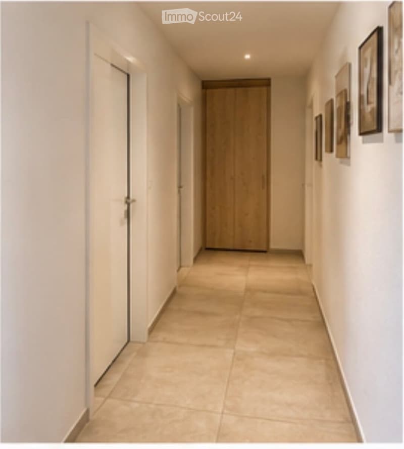 A clean, modern hallway with beige tiled flooring, white walls, and several doors. A wooden wardrobe is visible at the end of the hallway, and framed pictures are displayed on the right wall.