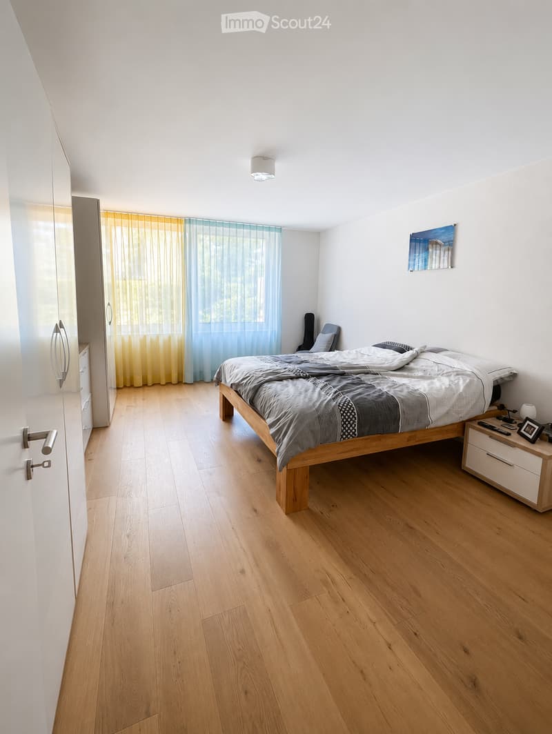 Bright bedroom with wooden flooring, a double bed with grey bedding, a nightstand with a lamp, a wardrobe, and a large window with sheer curtains.