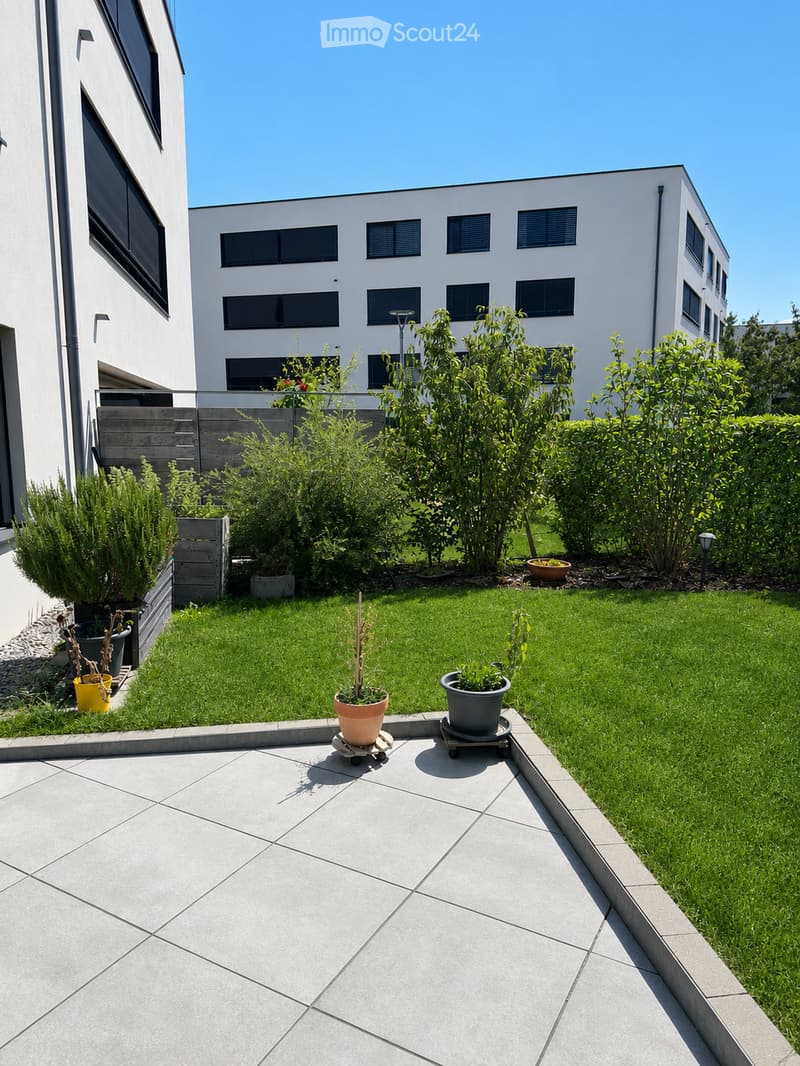 Well-maintained garden with green lawn, potted plants, and modern building in the background.