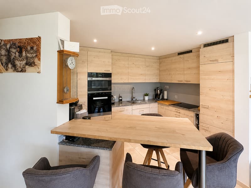 Modern kitchen with light wooden cabinets, granite countertops, and stainless steel appliances. Features a central wooden dining table with four grey chairs, a wall-mounted clock, and a picture of kittens on the wall.