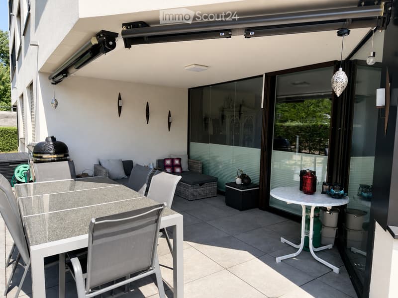 A modern outdoor terrace with a glass dining table and chairs, a sofa with cushions, a small ottoman, a stool with a teapot and cup, and a grill in the background.