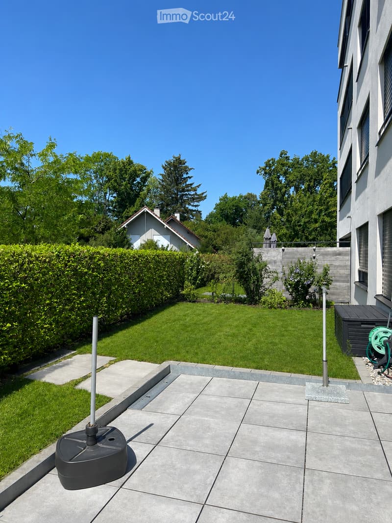 Well-maintained garden with a neatly trimmed lawn, concrete patio, and surrounding greenery. Includes a water feature and a small shed.