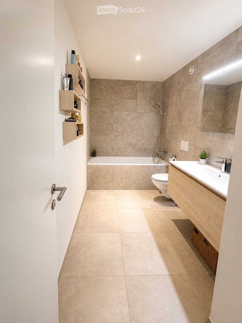 Modern bathroom with beige tiles, a bathtub, toilet, sink, wooden cabinet, shelves, and a large mirror.