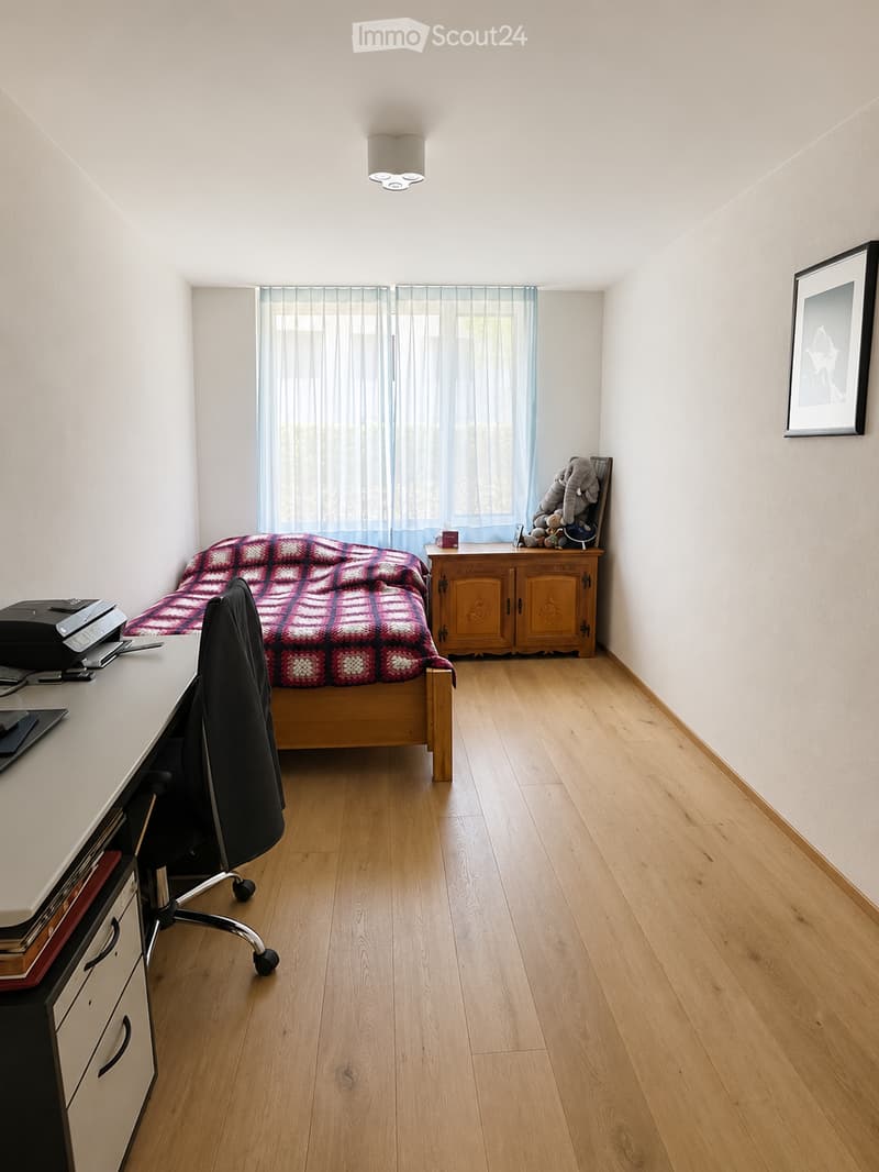 A well-lit bedroom with wooden flooring, a bed with a red and white checkered blanket, a desk with a chair, a cabinet with toys, and a framed picture on the wall.