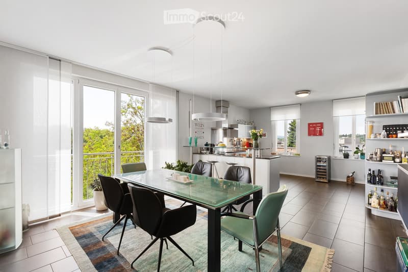 Modern open-plan dining and kitchen area with a glass dining table, black chairs, kitchen island, wine rack, shelves, and a view of greenery outside.
