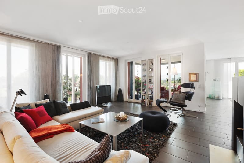 Modern living room with large windows, white walls, and a high ceiling. Features a white sectional sofa with colorful cushions, a coffee table, an ottoman, a sleek chair, a television, and a bookshelf with decorative items.