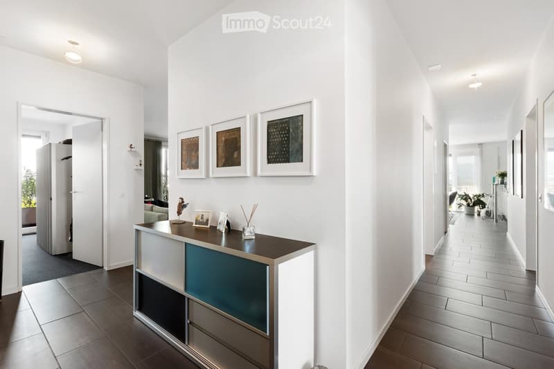 Modern hallway with white walls, tiled floors, and a reception desk with decorative items. The hallway features framed artwork and leads to other rooms.