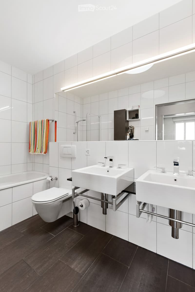 Modern bathroom with white tiles, dark wooden floor, double sink, toilet, bathtub, and a shower area. Features a large mirror, towel, and a small cabinet with toiletries.