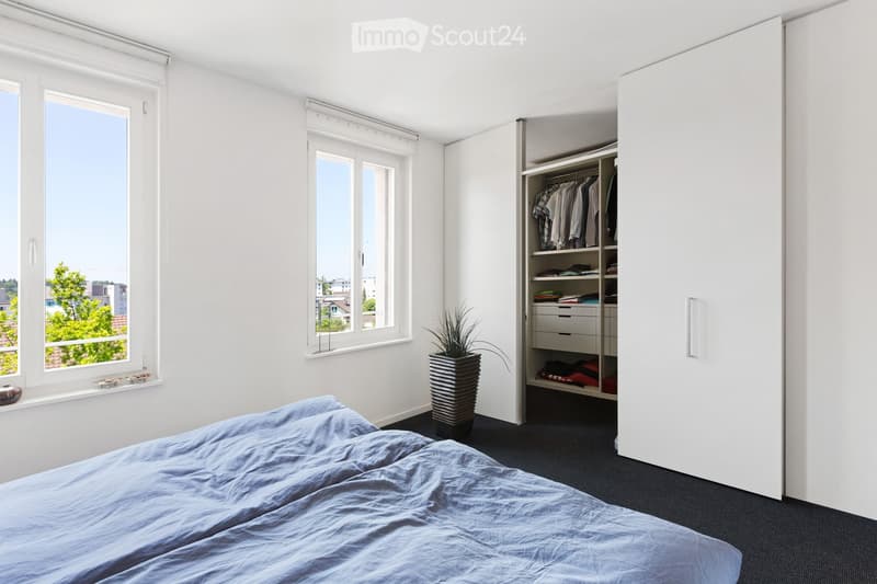 Modern bedroom with a neatly made bed, large windows with blinds, a tall wardrobe, and a potted plant on the floor.