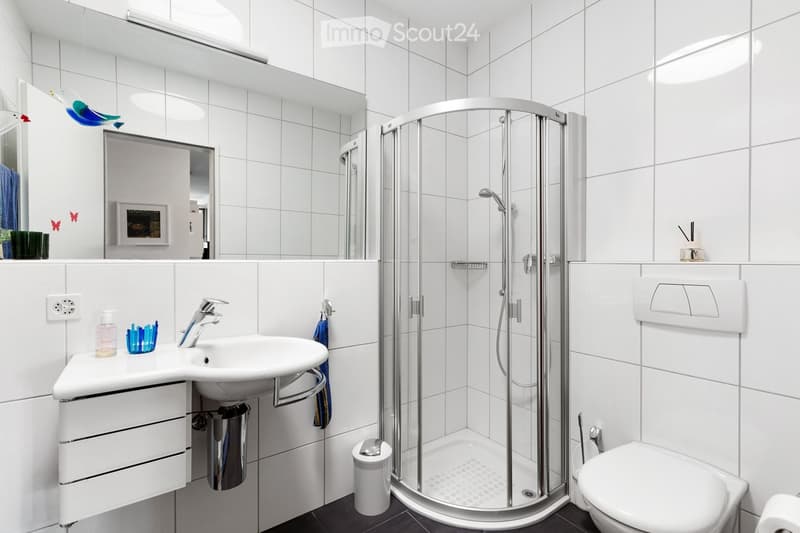 Modern bathroom with white tiled walls and floor, featuring a sink with a faucet, a mirror, a glass-enclosed shower, and a toilet. The sink has a small cabinet underneath, and there are decorative items on the counter.