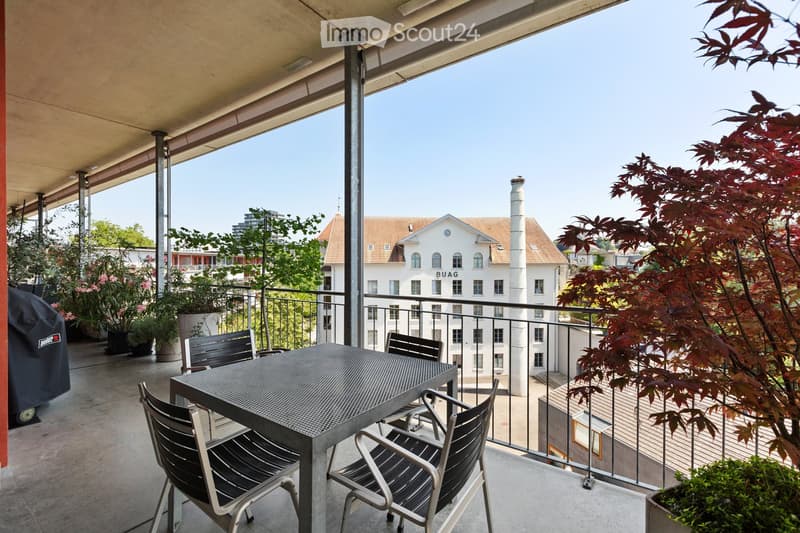 Balcony with a metal table and chairs, overlooking a cityscape with buildings and greenery.