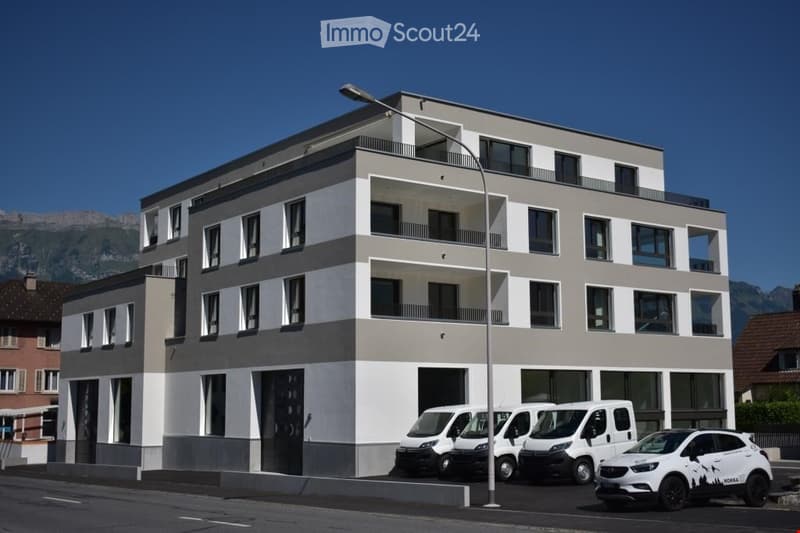 Modern multi-story building with white and grey facade, large windows, and balconies. Several vans are parked in front.