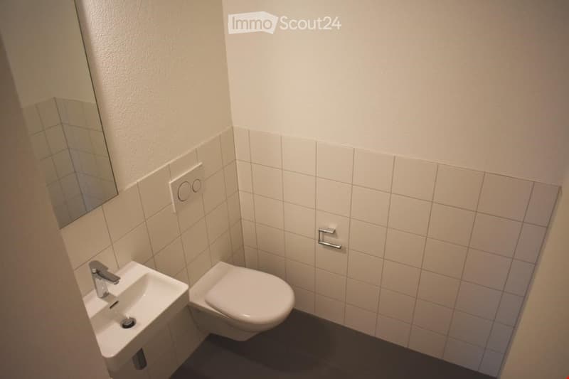 Modern bathroom with white tiled walls, a white sink, a toilet, and a mirror. The floor is dark grey.