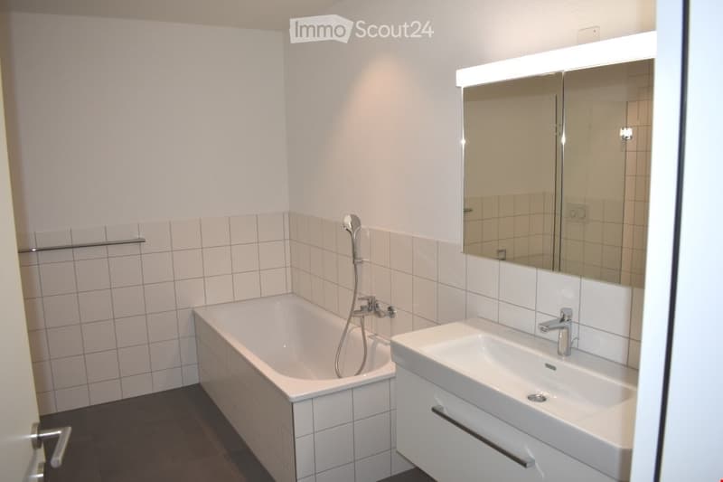Modern bathroom with a white bathtub, a white sink with a chrome faucet, a large mirror, and a showerhead attached to the wall.