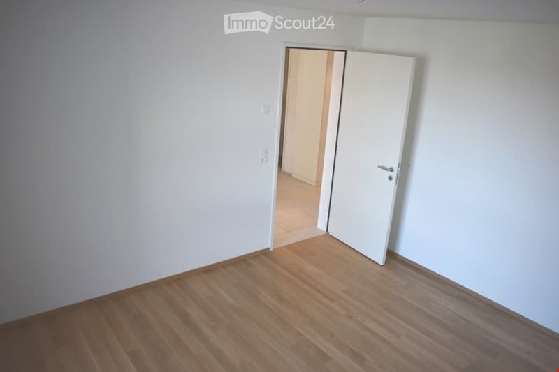 Empty room with light wooden flooring and white walls, featuring an open door leading to another room.