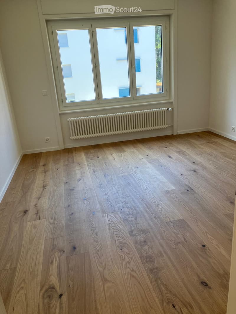 Empty room with wooden flooring, large window, radiator, and neutral-colored walls.