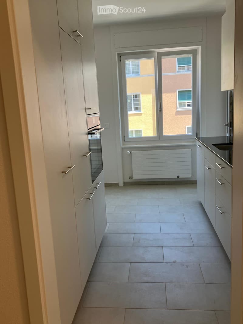 Empty kitchen with light-colored cabinets, tiled floor, and a window with a view of a neighboring building.