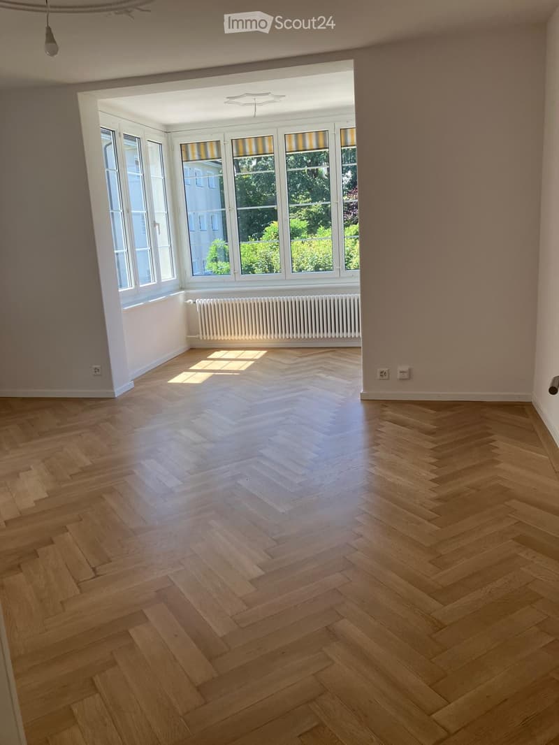 Empty room with light wooden herringbone flooring, large windows with striped blinds, radiator, and visible electrical outlets.