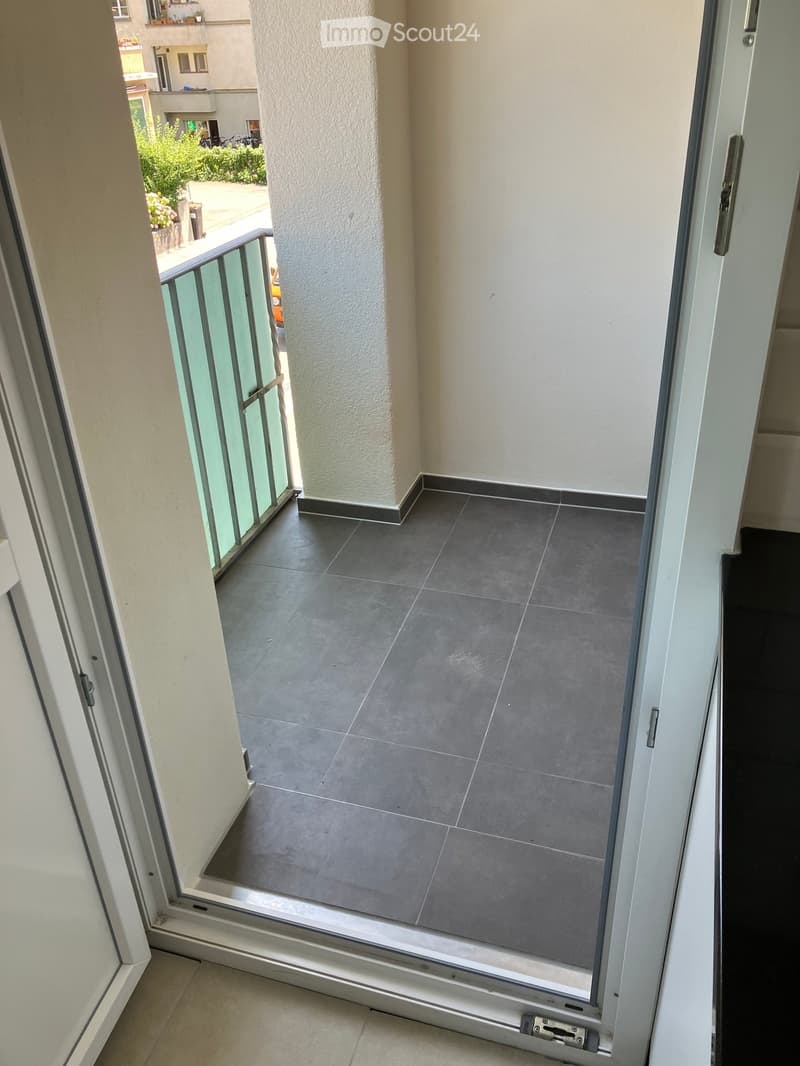 Balcony with grey tiled floor, glass railing, and open sliding door leading to the interior.