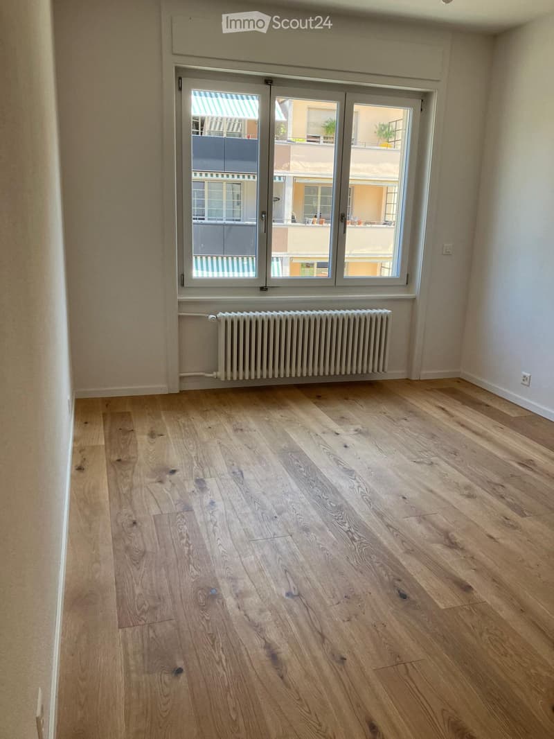 Empty room with light wooden flooring, white walls, a radiator, and large windows with a view of a balcony and neighboring buildings.
