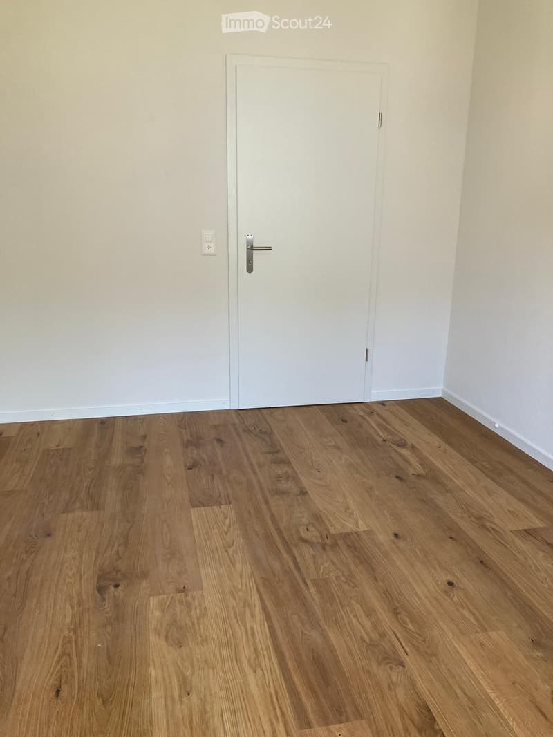 Empty room with light wooden flooring and white walls, featuring a closed white door and a power outlet on the wall.
