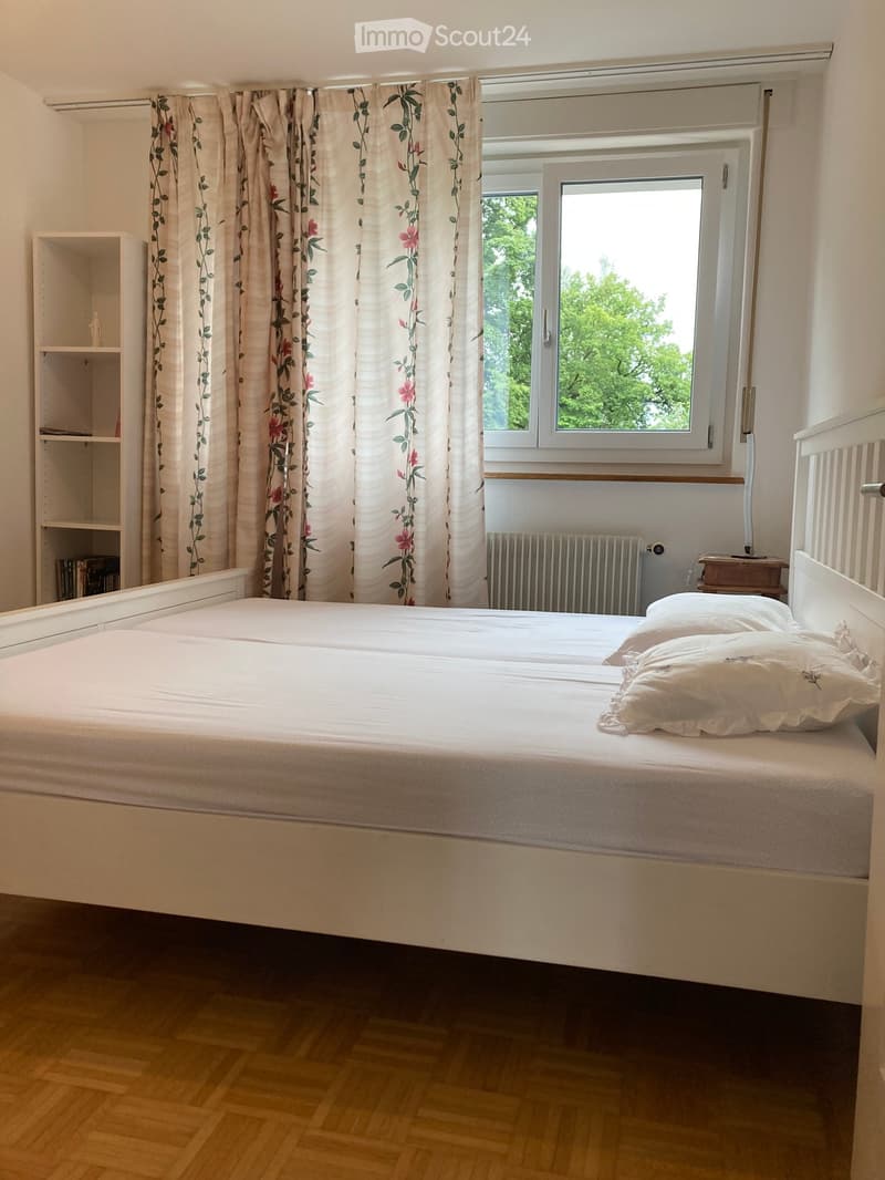 Cozy bedroom with a double bed, pillows, a nightstand, a shelf, and floral curtains. The room has wooden flooring and a window with a view of greenery.
