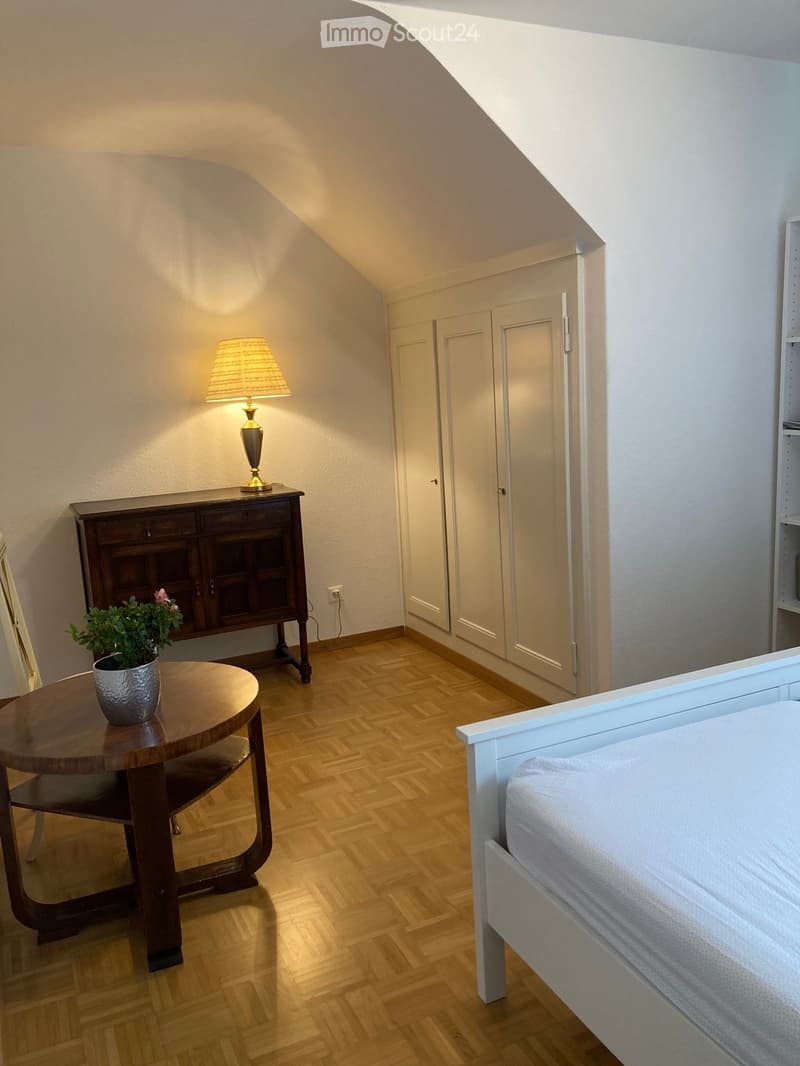 Cozy attic bedroom with wooden flooring, white walls, a bed, a small round table with a plant, a wooden cabinet, and a lamp on top.