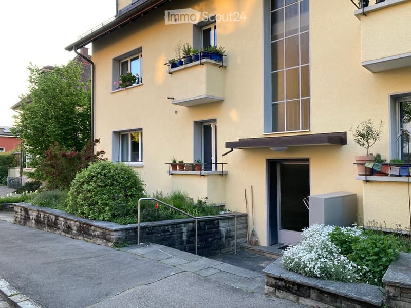 Two-story yellow building with multiple windows, balconies, and potted plants. Features a raised ground floor with a stone retaining wall and small garden area.