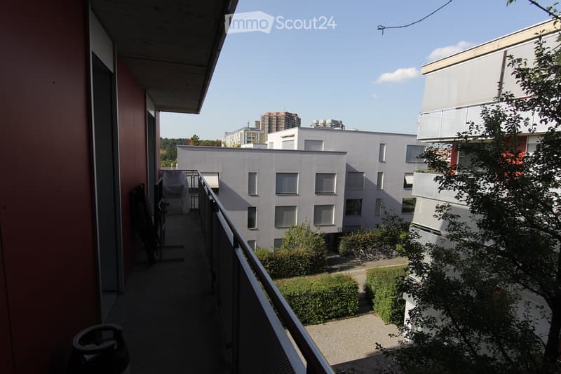 A balcony with a metal railing, a view of a neighboring building, and greenery