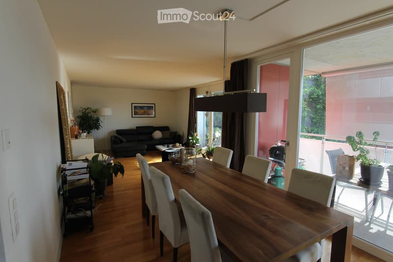 Dining room with a wooden dining table and chairs, white walls, sliding doors leading to a balcony with potted plants and a view of greenery outside