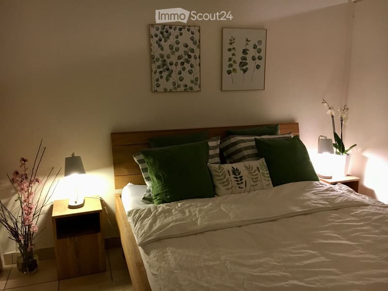 A neatly made bed with white sheets, a headboard, a nightstand with a lamp on both sides, two picture frames above the bed, a flower vase, and a small tree in a vase