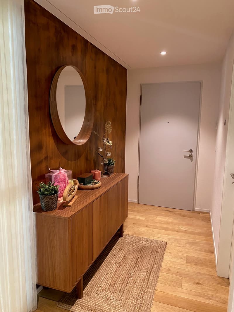Modern hallway with wooden floor, white walls, a round mirror, a small table with decorative items, and a closed door at the end.