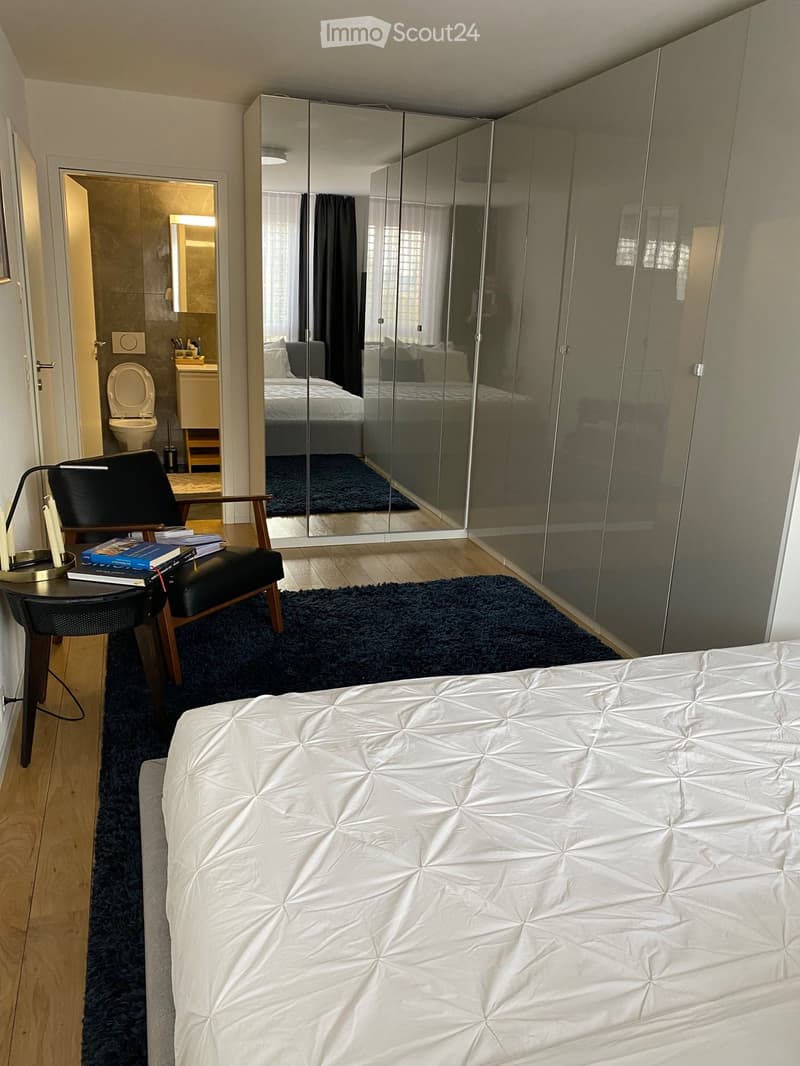 Modern bedroom with a large bed, a black chair, a small table with books and a lamp, a large mirror, and a view into an adjacent bathroom.