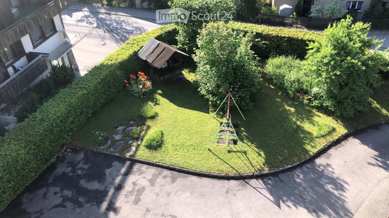 A small, well-maintained garden with a swing set, a small wooden shed, and neatly trimmed hedges. The garden is surrounded by a paved driveway and neighboring houses.