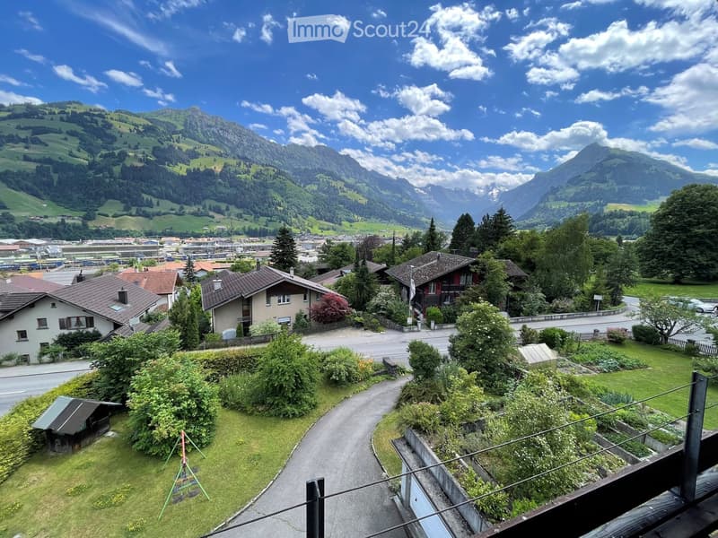 A scenic view from a balcony or terrace overlooking a town with houses, green hills, and distant mountains under a partly cloudy sky.