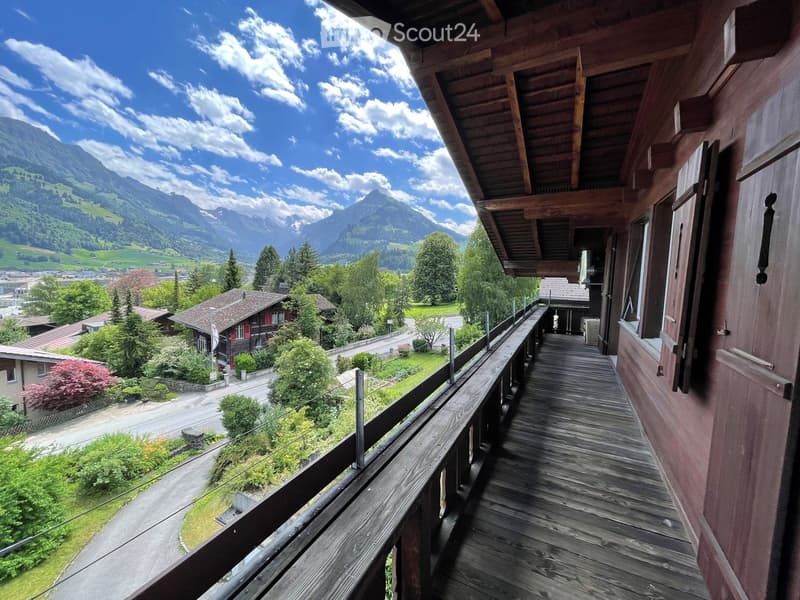 Wooden balcony with a railing, overlooking a scenic valley with mountains, trees, and houses.