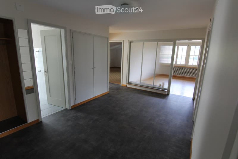 Empty room with grey carpeted floor, white walls, and multiple doors leading to other spaces. The room is completely unfurnished with no visible furniture or equipment.