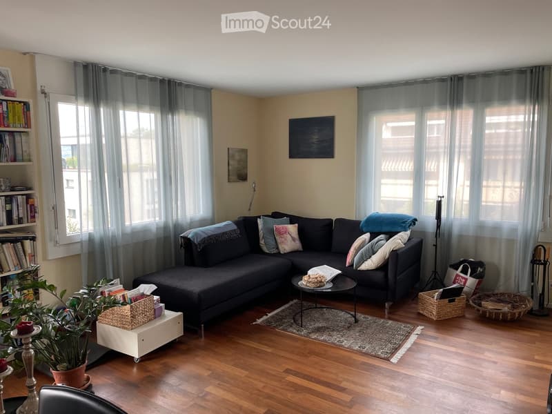 Modern living room with wooden flooring, large windows with grey curtains, a dark grey sectional sofa with multiple cushions, a small black coffee table, a white side table with a plant, and a bookshelf filled with books.