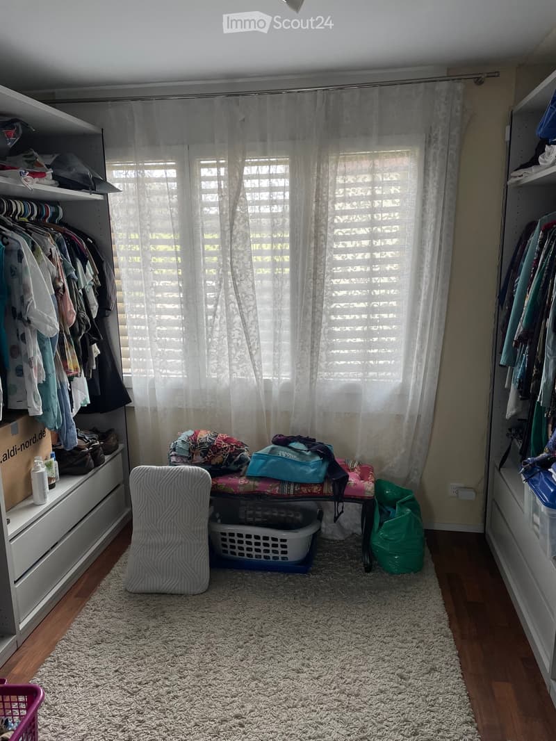 A well-lit room with wooden flooring, a white shaggy rug, a large window with blinds and sheer curtains, a white wardrobe with hanging clothes, a shelf with folded clothes and shoes, a small chair, a basket, and a stool with folded items on top.