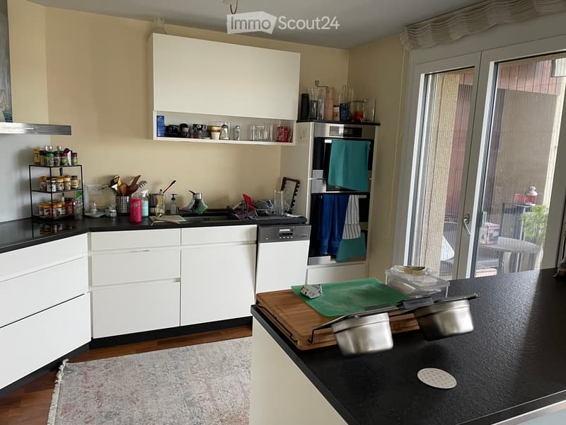 Modern kitchen with white cabinets, black countertops, and stainless steel appliances. Features a wooden cutting board with pots, a spice rack, and a dishwasher. A sliding glass door leads to a balcony.