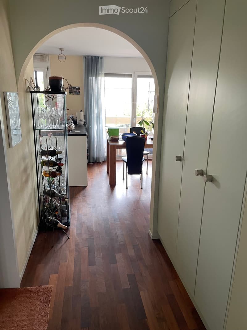 A well-lit hallway with wooden flooring, leading to a dining area with a table, chairs, and a wine rack on the left side.