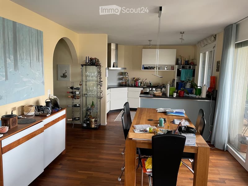 Bright kitchen with wooden flooring, white cabinets, and a central dining table with chairs. Includes a wine rack, sink, and various kitchen items on the counter.