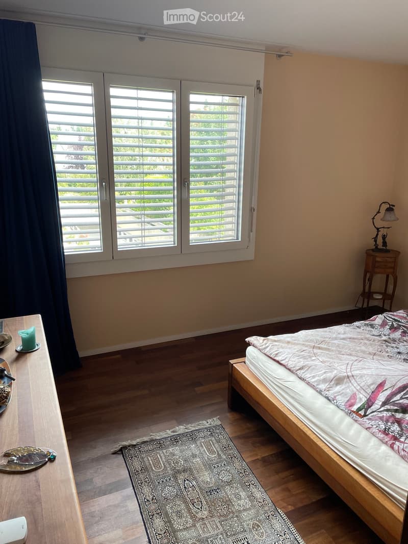 A well-lit bedroom with wooden flooring, a bed with a decorative quilt, a nightstand with a lamp, a small table with a candle, and a patterned rug.