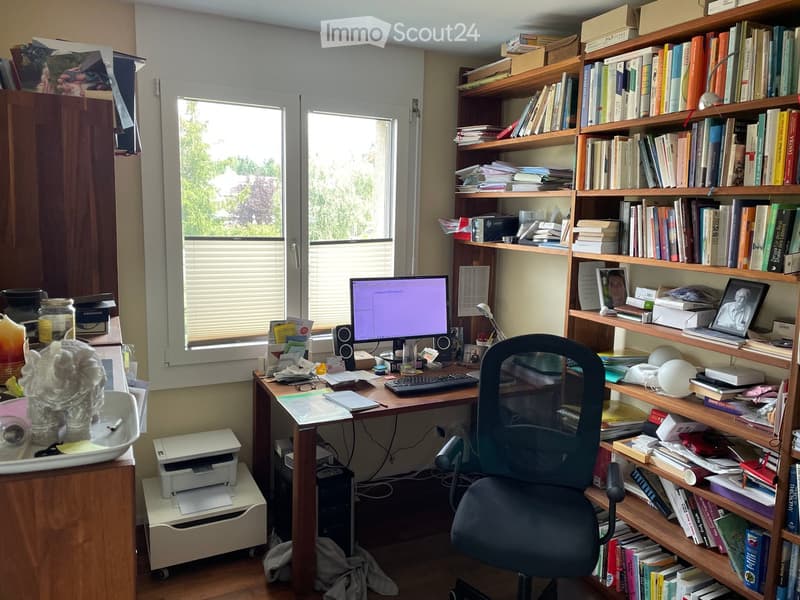 A well-organized home office with a wooden desk, computer setup, ergonomic chair, and a large bookshelf filled with books and decorative items.