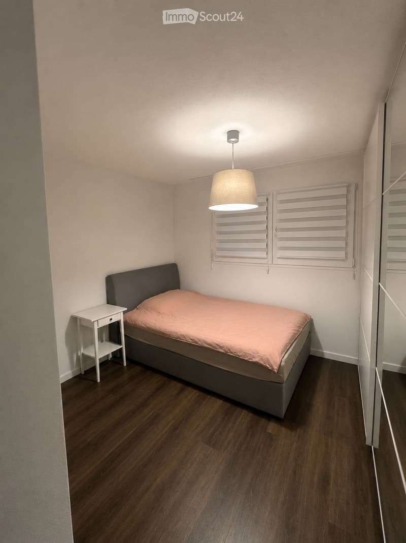 A small, modern bedroom with a single bed, a nightstand, and a hanging lamp. The room features wooden flooring and white walls with a window with shutters.