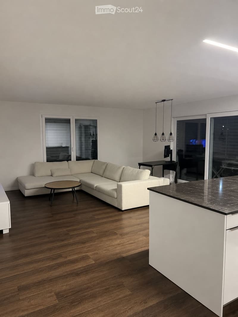 Modern living room with a white sectional sofa, a round coffee table, a kitchen island, and a small table with a screen. The room features wooden flooring and large windows with blinds.