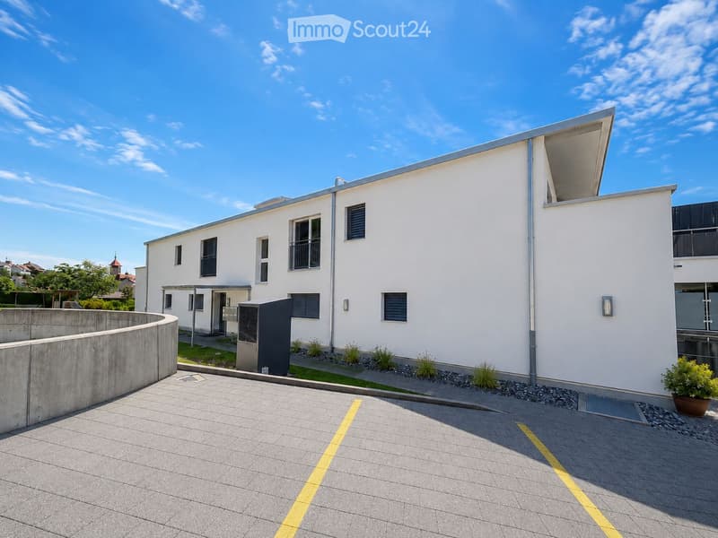 Modern two-story white building with large windows, a raised ground floor, and a small garden area.