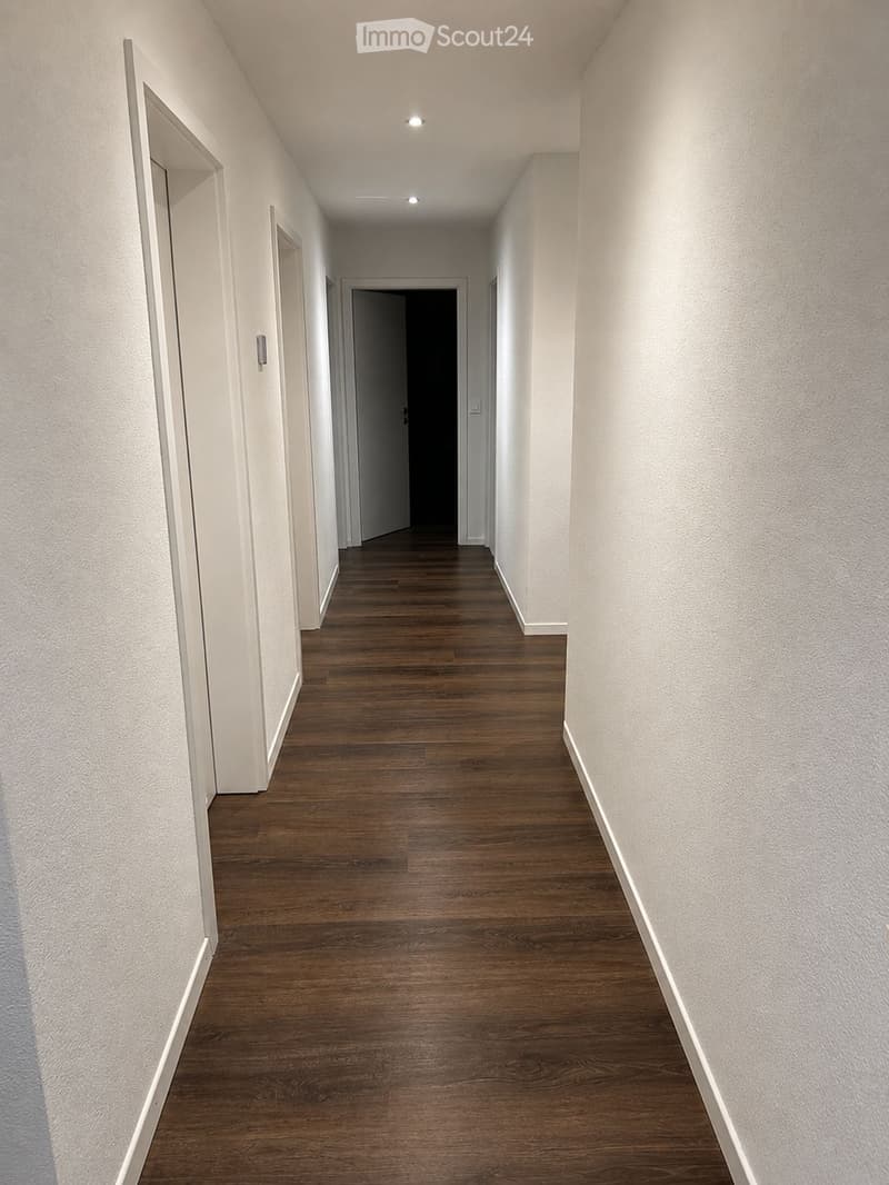 Empty hallway with wooden flooring, white walls, and recessed lighting. Doors are visible on both sides.