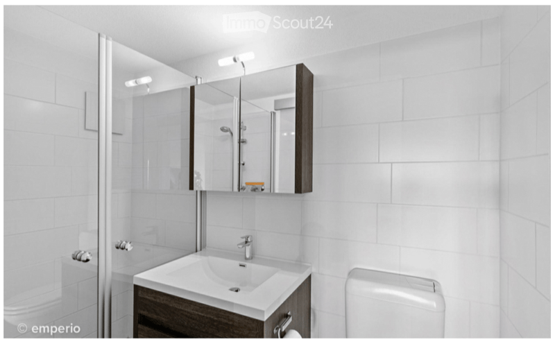 Modern bathroom with white tiles, a sink with a mirror cabinet, and a toilet. The shower area is visible in the background.